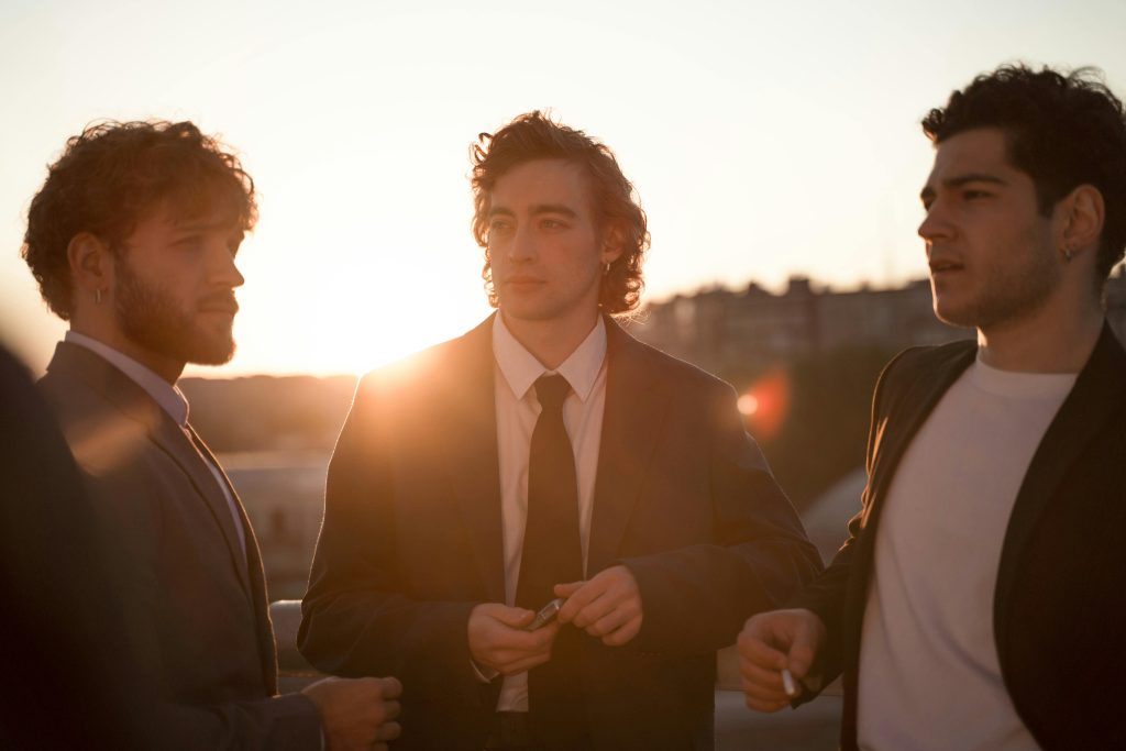 Three young men wearing suits, enjoying a stylish outdoor gathering during sunset, exuding a cool and relaxed vibe.