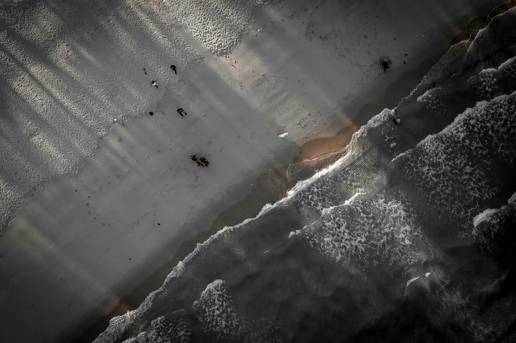 Drone view of waving sea washing sandy coast with people walking on beach and color sunlight illuminating seashore
